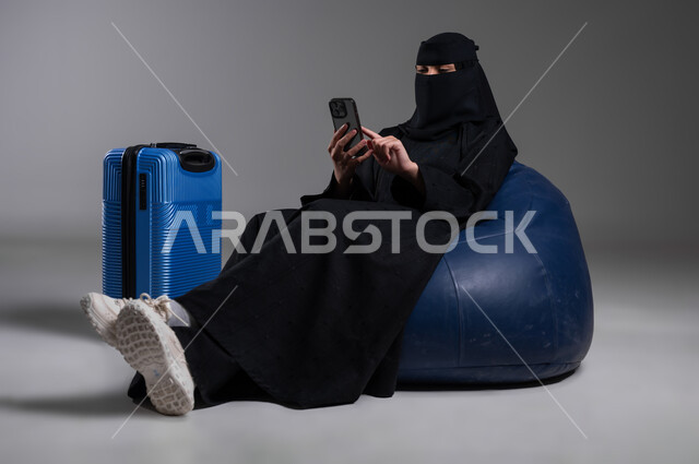 الذهاب في رحلات ممتعة، استخدام الهاتف المحمول في وقت الانتظار، الاستكشاف والمغامرات والاستجمام، الدردشة مع الأصدقاء وتصفح مواقع النت، بورتريه لامرأة عربية خليجية سعودية مسلمة منقبة ترتدي العباءة السوداء تجلس على كرسي مريح وبجانبها حقيبة سفر زرقاء، خلفية رمادية