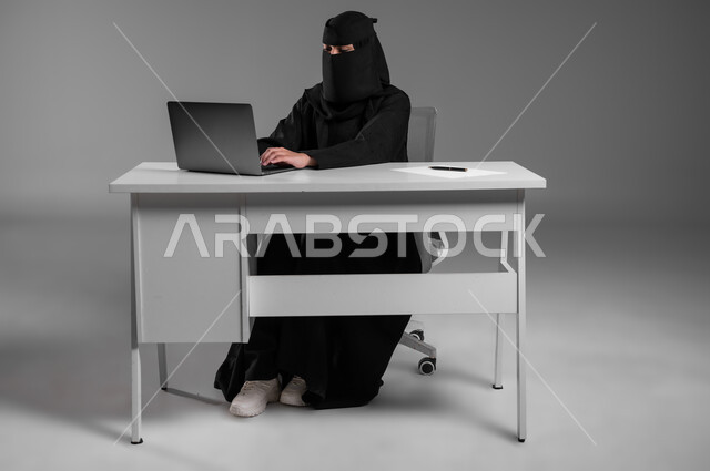 دمج التكنولوجيا والتقنيات الحديثة في المجال المكتبي، مهن ووظائف للنساء في السعودية، الإشراف والمتابعة على آلية سير العمل، بورتريه لامرأة عربية خليجية سعودية مسلمة منقبة ترتدي العباءة السوداء تجلس وراء المكتب وتعمل على الحاسوب المحمول، خلفية رمادية