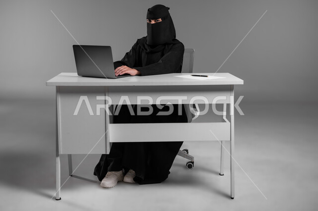 مهن ووظائف للنساء في السعودية، الإشراف والمتابعة على آلية سير العمل، استخدام التكنولوجيا والتقنيات الحديثة في المجال المكتبي، بورتريه لامرأة عربية خليجية سعودية مسلمة منقبة ترتدي العباءة السوداء تجلس وراء المكتب وتعمل على الحاسوب المحمول، خلفية رمادية