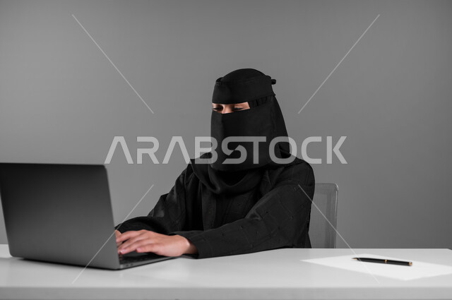 Supervising and following up on the workflow mechanism, using technology and modern techniques in the office field, a portrait of a veiled Saudi Gulf Arab Muslim woman wearing a black abaya sitting behind the desk and working on the laptop, professions and jobs for women in Saudi Arabia, gray background