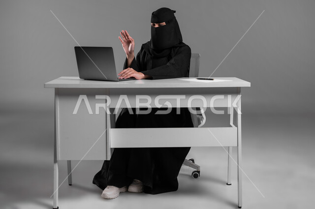 مفهوم العمل عن بعد، إنجاز المهام والأهداف المطلوبة،استخدام التكنولوجيا والتقنيات الحديثة في المجال المكتبي، بورتريه لامرأة عربية خليجية سعودية مسلمة منقبة ترتدي العباءة السوداء تجلس وراء المكتب تجري مكالمة فيديو، مهن ووظائف للنساء في السعودية، خلفية رمادية