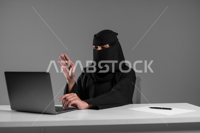 إنجاز المهام والأهداف المطلوبة،استخدام التكنولوجيا والتقنيات الحديثة في المجال المكتبي، مفهوم العمل عن بعد، بورتريه لامرأة عربية خليجية سعودية مسلمة منقبة ترتدي العباءة السوداء تجلس وراء المكتب تجري مكالمة فيديو، مهن ووظائف للنساء في السعودية، خلفية رمادية