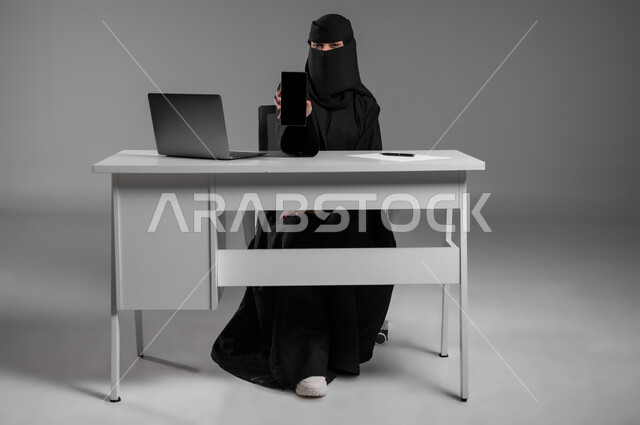 استخدام التكنولوجيا الحديثة في المهن المكتبية، عرض شاشة سوداء فارغة، متابعة العمل عبر الحاسوب المحمول، بورتريه لامرأة عربية خليجية سعودية مسلمة منقبة ترتدي العباءة السوداء تجلس وراء المكتب تمسك بيدها جهاز الجوال، وظائف للنساء في السعودية، خلفية رمادية