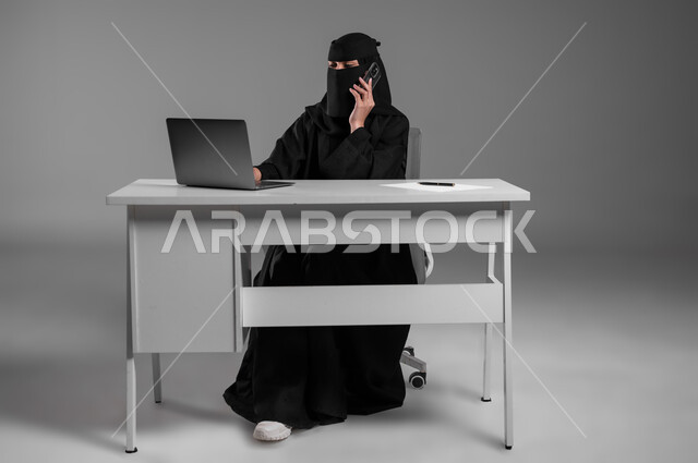 الاستشارات وتدوين الملاحظات، متابعة العمل عبر الحاسوب المحمول، بورتريه لامرأة عربية خليجية سعودية مسلمة منقبة ترتدي العباءة السوداء تجلس وراء المكتب تمسك بيدها جهاز الجوال، استخدام التكنولوجيا الحديثة في المهن المكتبية، وظائف للنساء في السعودية، خلفية رمادية
