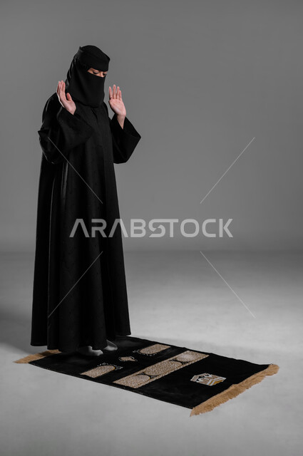 الوقوف على السجادة استعداداً للصلاة، الالتزام بتعاليم الدين الإسلامي، بورتريه لامرأة عربية خليجية سعودية مسلمة منقبة ترتدي العباءة السوداء ترفع يديها وتكبر لتبدأ صلاتها، صورة الجسم بالطول الكامل، مفهوم الحشمة والحياء، خلفية رمادية