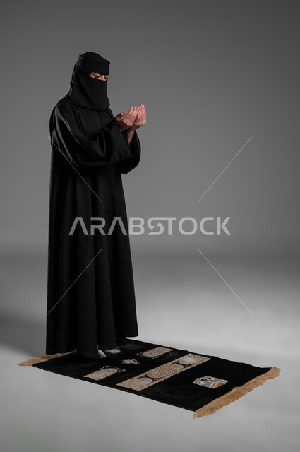 رفع الكفين والتوجه إلى بابه الكريم، أداء الصلوات والفرائض في أوقاتها، الالتزام بتعاليم الدين الإسلامي، بورتريه لامرأة عربية خليجية سعودية مسلمة منقبة ترتدي العباءة السوداء تفرش سجادة الصلاة أمامها وتدعو في صلاتها، صورة الجسم بالطول الكامل، خلفية رمادية