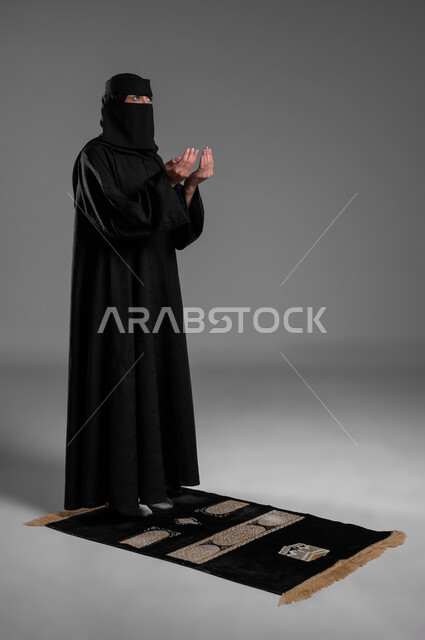 الدعاء والقيام بالطاعات والعبادات،أداء الصلوات والفرائض في أوقاتها، الالتزام بتعاليم الدين الإسلامي، بورتريه لامرأة عربية خليجية سعودية مسلمة منقبة ترتدي العباءة السوداء تفرش سجادة الصلاة أمامها وتدعو في صلاتها، صورة الجسم بالطول الكامل، خلفية رمادية