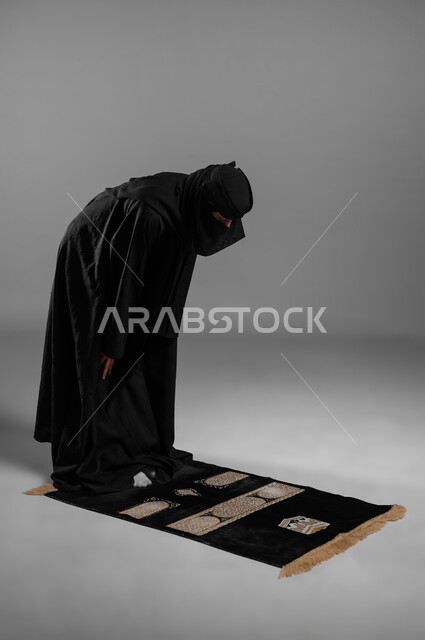القيام بالطاعات والعبادات وترديد الأذكار،أداء الصلوات والفرائض في أوقاتها، الالتزام بتعاليم الدين الإسلامي، بورتريه لامرأة عربية خليجية سعودية مسلمة منقبة ترتدي العباءة السوداء تفرش سجادة الصلاة أمامها وتركع في صلاتها، صورة الجسم بالطول الكامل، خلفية رمادية