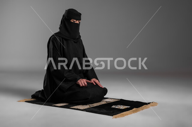 المحافظة على الصلوات والفرائض في أوقاتها، الالتزام بتعاليم الدين الإسلامي، ترديد الأذكار وأداء الطاعات، بورتريه لامرأة عربية خليجية سعودية مسلمة منقبة ترتدي العباءة السوداء تجلس على سجادة الصلاة وتصلي، خلفية رمادية