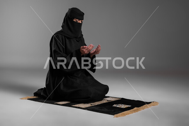 التقرب من الله بالخضوع  والدعاء، الحفاظ على الصلوات والفرائض، الالتزام بتعاليم الدين الإسلامي، ترديد الأذكار وأداء الطاعات، بورتريه لامرأة عربية خليجية سعودية مسلمة منقبة ترتدي العباءة السوداء تجلس على سجادة الصلاة وتفتح كفيها وتدعو الله، خلفية رمادية