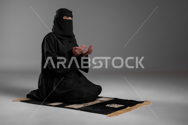 الخضوع  بالدعاء والتقرب من الله، الحفاظ على الصلوات والفرائض، الالتزام بتعاليم الدين الإسلامي، ترديد الأذكار وأداء الطاعات، بورتريه لامرأة عربية خليجية سعودية مسلمة منقبة ترتدي العباءة السوداء تجلس على سجادة الصلاة وتفتح كفيها وتدعو الله، خلفية رمادية