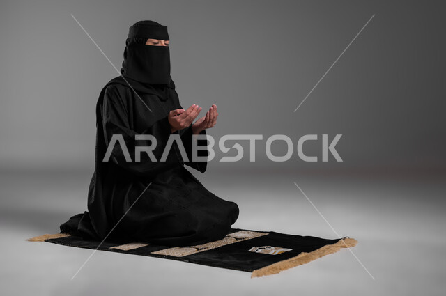 الدعاء والخضوع والتقرب من الله، الحفاظ على الصلوات والفرائض، الالتزام بتعاليم الدين الإسلامي، ترديد الأذكار وأداء الطاعات، بورتريه لامرأة عربية خليجية سعودية مسلمة منقبة ترتدي العباءة السوداء تجلس على سجادة الصلاة وتفتح كفيها وتدعو الله، خلفية رمادية