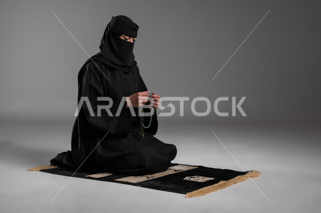 الاستغفار وذكر الله والإمساك بالسبحة، الحفاظ على الصلوات والفرائض، الالتزام بتعاليم الدين الإسلامي، ترديد الأذكار وأداء الطاعات، بورتريه لامرأة عربية خليجية سعودية مسلمة منقبة ترتدي العباءة السوداء تجلس على سجادة الصلاة وتقوم بالتسبيح، خلفية رمادية