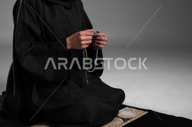 الإمساك بالسبحة والاستغفار وذكر الله، الحفاظ على الصلوات والفرائض، الالتزام بتعاليم الدين الإسلامي، ترديد الأذكار وأداء الطاعات، بورتريه مقرب لامرأة عربية خليجية سعودية مسلمة ترتدي العباءة السوداء تجلس على سجادة الصلاة وتقوم بالتسبيح، خلفية رمادية