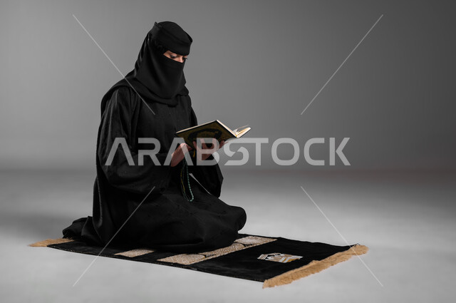 مفهوم الحشمة والالتزام بتعاليم الدين الإسلامي، ترديد الأذكار والتسبيح والاستغفار بعد الصلوات، قراءة القرآن الكريم وأداء الطاعات، بورتريه لامرأة عربية خليجية سعودية مسلمة منقبة ترتدي العباءة السوداء تجلس على سجادة الصلاة وتمسك كتاب الله، خلفية رمادية