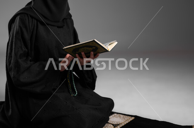 الحفاظ على الصلوات والفرائض، الالتزام بتعاليم الدين الإسلامي، ترديد الأذكار والتسبيح والاستغفار، قراءة القرآن الكريم وأداء الطاعات، بورتريه مقرب لامرأة عربية خليجية سعودية مسلمة ترتدي العباءة السوداء تجلس على سجادة الصلاة وتمسك كتاب الله، خلفية رمادية