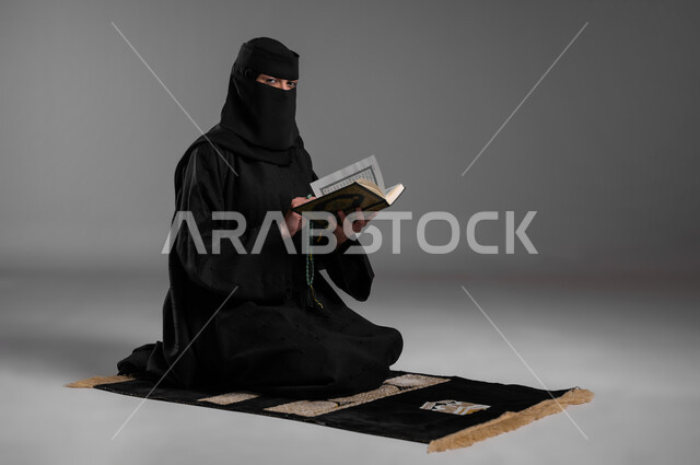 التسبيح والاستغفار وترديد الأذكار بعد الصلوات، قراءة القرآن الكريم وأداء الطاعات، بورتريه لامرأة عربية خليجية سعودية مسلمة منقبة ترتدي العباءة السوداء تجلس على سجادة الصلاة وتمسك كتاب الله، مفهوم الحشمة والالتزام بتعاليم الدين الإسلامي، خلفية رمادية