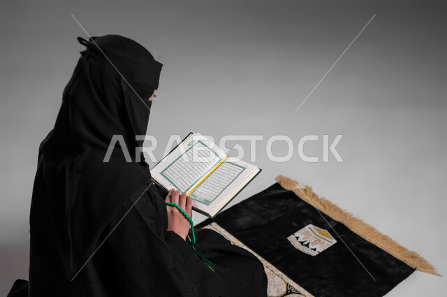 الالتزام بتعاليم الدين الإسلامي ومفهوم الحشمة، ترديد الأذكار والتسبيح والاستغفار بعد الصلوات، قراءة القرآن الكريم وأداء الطاعات، بورتريه من الخلف لامرأة عربية خليجية سعودية مسلمة منقبة ترتدي العباءة السوداء تجلس على سجادة الصلاة وتمسك كتاب الله، خلفية رمادية