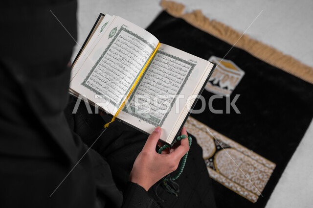 قراءة القرآن الكريم وأداء الطاعات، بورتريه من الأعلى لامرأة عربية خليجية سعودية مسلمة منقبة ترتدي العباءة السوداء تجلس على سجادة الصلاة وتمسك كتاب الله، التسبيح والاستغفار وترديد الأذكار بعد الصلوات، مفهوم الحشمة والالتزام بتعاليم الدين الإسلامي، خلفية رمادية