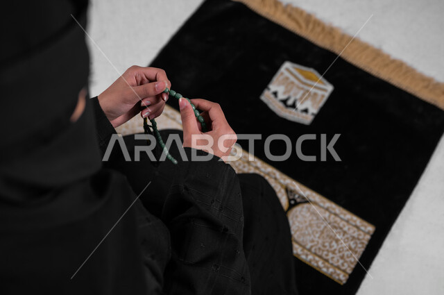 أداء الطاعات والتقرب من الله، بورتريه من الأعلى لامرأة عربية خليجية سعودية مسلمة منقبة ترتدي العباءة السوداء تجلس على سجادة الصلاة وتمسك السبحة بيدها، التسبيح والاستغفار وقراءة الأذكار بعد الصلوات، مفهوم الحشمة والالتزام بتعاليم الدين الإسلامي، خلفية رمادية