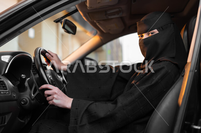 الجلوس خلف المقود والنظر للكاميرا بثقة, الانضباط بقواعد السلامة وربط حزام الأمان، الاعتماد على النفس في التنقلات عند النساء، حرية القيادة للمرأة السعودية، صورة من الجانب لسيدة عربية خليجية سعودية مسلمة منقبة ترتدي العباءة السوداء وتقود في الطرق العامة بمهارة