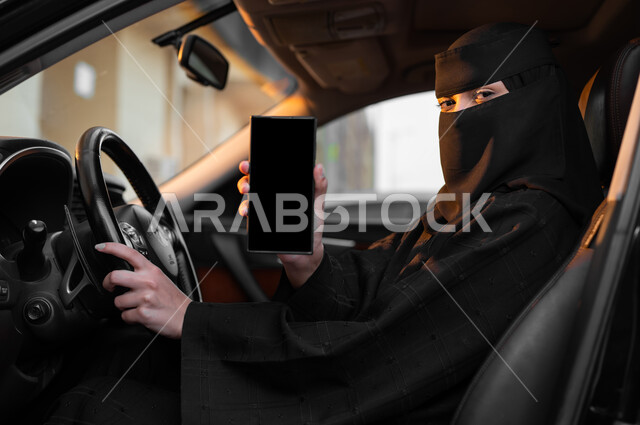 النظر للكاميرا والإمساك بالهاتف المحمول مع عرض شاشة سوداء فارغة, اعتماد النساء على أنفسهن في وسائل النقل، حرية قيادة السيارة للمرأة السعودية، سيدة عربية خليجية سعودية مسلمة منقبة ترتدي العباءة التقليدية تجلس داخل سيارتها، مفهوم الحشمة والالتزام