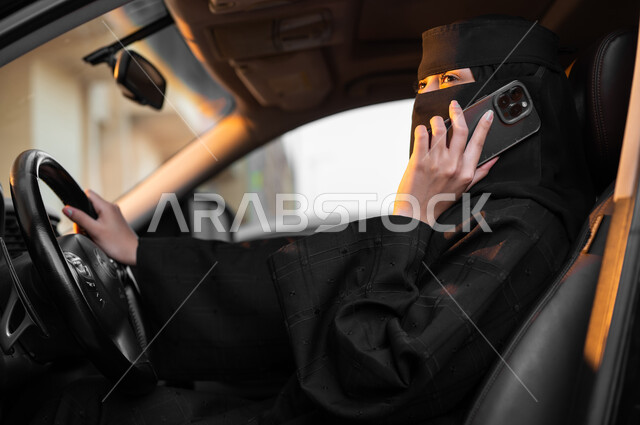 الإمساك بالهاتف المحمول والتحدث به, حرية قيادة السيارة للمرأة السعودية، صورة من الجانب لسيدة عربية خليجية سعودية مسلمة منقبة ترتدي العباءة السوداء تجلس داخل سيارتها وتتقوم بمكالمة هاتفية مهمة، مفهوم الحشمة والالتزام, الاعتماد على النفس في وسائل النقل