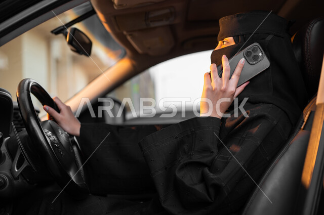 Making important phone calls using a mobile phone, the freedom to drive a car for Saudi women, a picture from the side of a veiled Saudi Gulf Arab Muslim woman wearing a black abaya sitting inside her car and holding a mobile phone, the concept of decency and commitment, self-reliance in transportation