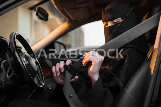 Discipline with safety rules, preparing the car before setting off, freedom to drive for Saudi women, a close-up photo from the side of a veiled Saudi Gulf Arab woman wearing a black abaya sitting inside her car and fastening her seat belt, the concept of decency and commitment, self-reliance in transportation