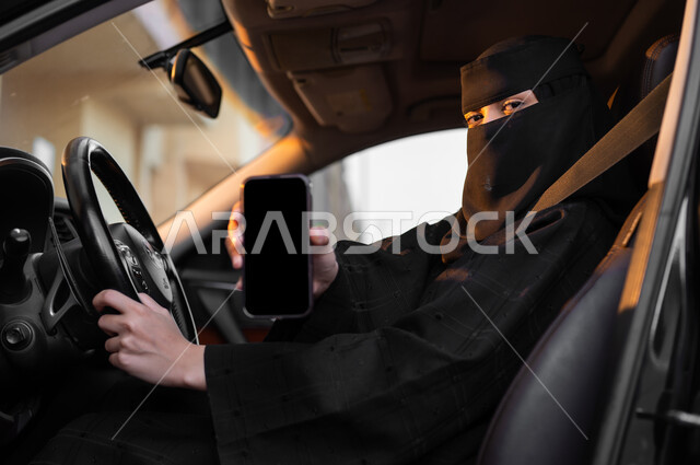 الإمساك بالهاتف المحمول وعرض شاشة سوداء فارغة, اعتماد النساء على أنفسهن في وسائل النقل، حرية قيادة السيارة للمرأة السعودية، سيدة عربية خليجية سعودية مسلمة منقبة ترتدي العباءة التقليدية تجلس داخل سيارتها، مفهوم الحشمة والالتزام، الاستفادة من التقنيات الحديثة