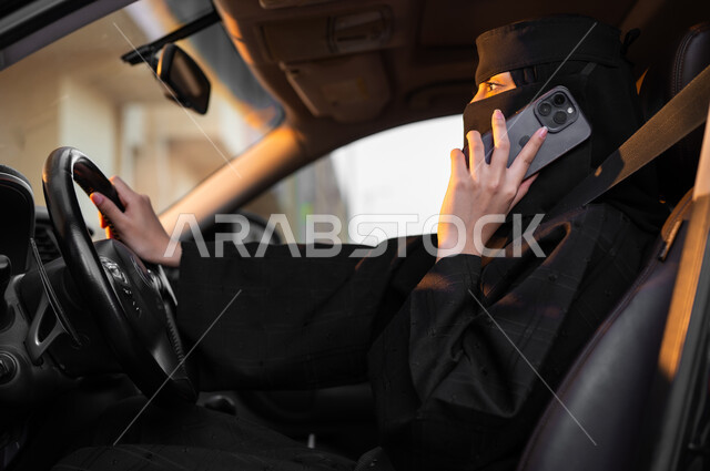 إجراء المكالمات الهاتفية باستخدام الجوال, حرية قيادة السيارة للمرأة السعودية، صورة من الجانب لسيدة عربية خليجية سعودية مسلمة منقبة ترتدي العباءة السوداء تجلس داخل سيارتها وتمسك الهاتف المحمول، مفهوم الحشمة والالتزام, الاعتماد على النفس في وسائل النقل