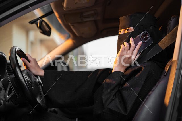 استخدام الجوال في إجراء المكالمات الهاتفية, حرية قيادة السيارة للمرأة السعودية، صورة من الجانب لسيدة عربية خليجية سعودية مسلمة منقبة ترتدي العباءة السوداء تجلس داخل سيارتها وتمسك الهاتف المحمول، مفهوم الحشمة والالتزام, الاعتماد على النفس في وسائل النقل
