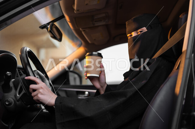 الحصول على المشروب المفضل قبل الانطلاق, حرية قيادة السيارة للمرأة السعودية، سيدة عربية خليجية سعودية مسلمة منقبة ترتدي العباءة التقليدية تجلس داخل سيارتها وتمسك بيدها كوب ورقي، مفهوم الحشمة والالتزام, اعتماد النساء على أنفسهن في وسائل النقل والمواصلات