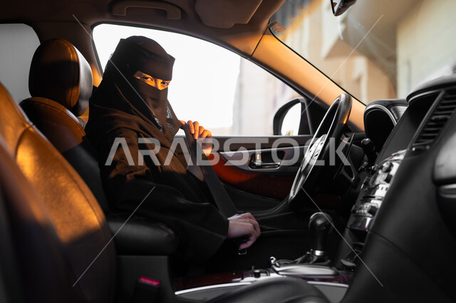 تجهيز السيارة قبل الانطلاق, حرية القيادة للمرأة السعودية، صورة من الجانب لسيدة عربية خليجية سعودية مسلمة منقبة ترتدي العباءة السوداء تجلس داخل سيارتها وتقوم بربط حزام الأمان، مفهوم الحشمة والالتزام, الاعتماد على النفس في التنقلات، اتباع قوانين السلامة