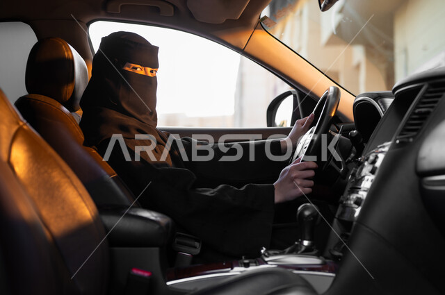 الاعتماد على الذات في التنقلات والمواصلات، مفهوم الحشمة والالتزام, حرية قيادة السيارة للمرأة السعودية، صورة من الجانب لسيدة عربية خليجية سعودية مسلمة منقبة ترتدي العباءة السوداء تجلس داخل سيارتها وتضع يديها على المقود، النظر للكاميرا بإيماءات الثقة بالنفس