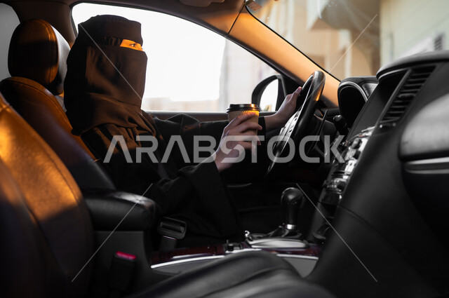 الحصول على المشروبات الساخنة, حرية قيادة السيارة للمرأة السعودية، صورة من الجانب لسيدة عربية خليجية سعودية مسلمة منقبة ترتدي العباءة السوداء تجلس داخل سيارتها وتحمل بيدها كوب من القهوة، مفهوم الحشمة والالتزام, اعتماد النساء على أنفسهن في وسائل النقل والمواصلات