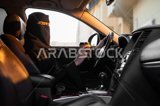 الحصول على كوب من القهوة, حرية قيادة السيارة للمرأة السعودية، صورة من الجانب لسيدة عربية خليجية سعودية مسلمة منقبة ترتدي العباءة السوداء تجلس داخل سيارتها وتحمل مشروبها المفضل بيدها، مفهوم الحشمة والالتزام, اعتماد النساء على أنفسهن في وسائل النقل والمواصلات