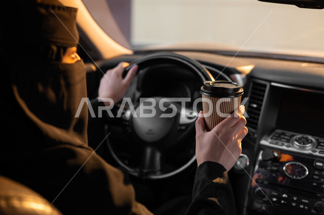 الجلوس داخل السيارة والإمساك بكوب ورقي, حرية قيادة السيارة للمرأة السعودية، صورة من الجانب لسيدة عربية خليجية سعودية مسلمة منقبة ترتدي العباءة السوداء التقليدية تمسك مشروبها المفضل، مفهوم الحشمة والالتزام, اعتماد النساء على أنفسهن في وسائل النقل والمواصلات