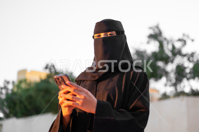 استخدام التقنيات الحديثة والاستفادة منها, امرأة عربية خليجية سعودية مسلمة منقبة ترتدي العباءة السوداء التقليدية تمسك الهاتف المحمول بيدها، التواصل  والدردشة مع الأهل والأصدقاء, مفهوم الحشمة والالتزام, الأناقة والاهتمام بالمظهر الخارجي، خلفية الطبيعة