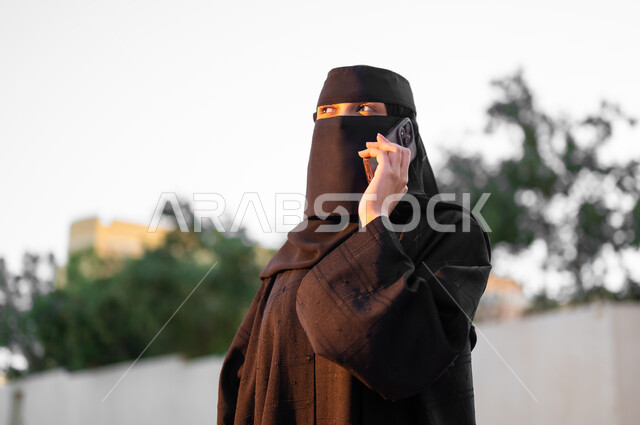 إجراء مكالمة صوتية باستخدام الهاتف المحمول, امرأة عربية خليجية سعودية مسلمة منقبة ترتدي العباءة السوداء التقليدية تقوم بالتواصل مع الأهل والأصدقاء, مفهوم الحشمة والالتزام, الوقوف باستقامة بإيماءات الثقة بالنفس، الأناقة والاهتمام بالمظهر الخارجي، خلفية الطبيعة