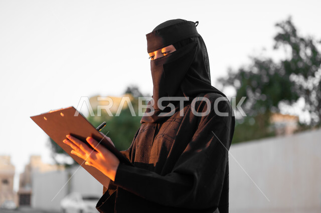 Holding a paper document and taking notes, a veiled Saudi Gulf Arab Muslim woman wearing the traditional black abaya, writing and recording important information, the concept of modesty and commitment, standing upright with gestures of self-confidence, elegance and attention to external appearance, nature background