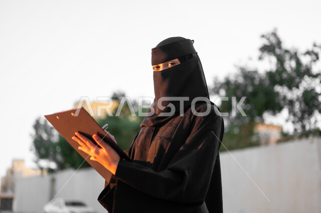 تدوين وكتابة الملاحظات, امرأة عربية خليجية سعودية مسلمة منقبة ترتدي العباءة السوداء التقليدية تقف وتمسك بيدها مستند ورقي, النظر إلى الكاميرا بتعبيرات الجدية, مفهوم الحشمة والالتزام, الأناقة والاهتمام بالمظهر الخارجي، خلفية الطبيعة