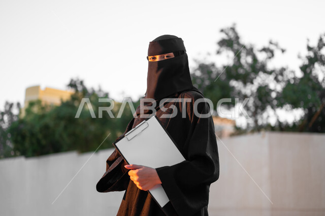 الوقوف بإيماءات الثقة بالنفس, امرأة عربية خليجية سعودية مسلمة منقبة ترتدي العباءة السوداء التقليدية تمسك بيدها مستند ورقي, النظر إلى الكاميرا بتعبيرات الجدية, مفهوم الحشمة والالتزام, الأناقة والاهتمام بالمظهر الخارجي، خلفية الطبيعة