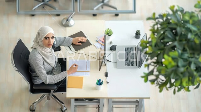 التوظيف في مجال خدمة العملاء والرد على الاسئلة والاستفسارات, تمكين عمل المرأة في المجتمع السعودي, صورة من الأعلى لشابة عربية خليجية سعودية محجبة, الجلوس وراء المكتب وتنفيذ الأعمال على جهاز الحاسوب,  مهن و وظائف نسائية سعودية