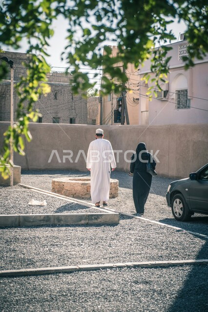 مفهوم الرابطة الزوجية والألفة بين الأزواج, صورة من الخلف لزوجين عربيين خلييجين عمانيين يمشيان في الحارات الشعبية القديمة, قضاء الأوقات برفقة العائلة, خلفية البيوت والشقق السكنية على الطراز المعماري القديم