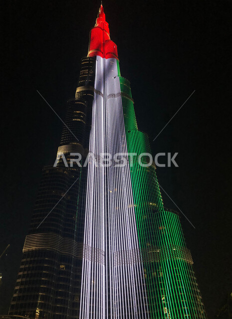 صورة مقربة لبرج خلفية المضاء بعلم دولة الامارات في وقت الليل، الاحتفال باليوم الوطني الاماراتي، التقدم والتطور المعماري في البلاد، معالم إمارة دبي السياحية