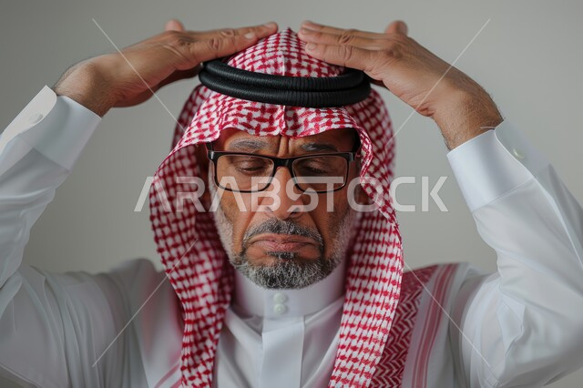 التفاجؤ والاستغراب من شيء ما, خسارة وفقدان شيء معين, الوقوف بإيماءات التعجب والدهشة، بورتريه مقرب لرجل كبير السن عربي خليجي سعودي يرتدي الشماغ والثوب التقليدي والنظارة الطبية يضع يديه على رأسه بتعابير الصدمة والذهول، خلفية رمادية