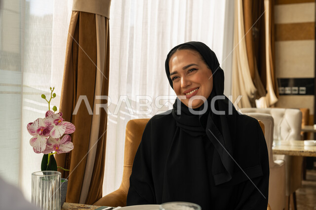 قضاء الأوقات الممتعة في الخارج, إيماءات السعادة و السرور, الاستمتاع بالأجواء المميزة في المطعم, امراة عربية خليجية إماراتية مبتسمة محجبة ترتدي العباءة السوداء تجلس في إحدى المطاعم في الإمارات, أنشطة ترفيهية مسلية