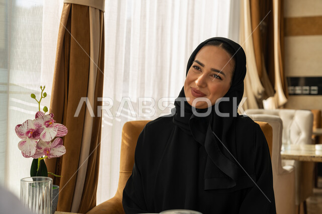 الاستمتاع بالأجواء المميزة في المطعم, قضاء الأوقات الممتعة في الخارج, إيماءات السعادة و السرور, امراة عربية خليجية إماراتية مبتسمة محجبة ترتدي العباءة السوداء تجلس في إحدى المطاعم في الإمارات, أنشطة ترفيهية مسلية