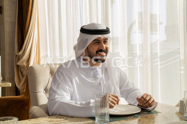 الاستمتاع بالأجواء المميزة في المطعم, قضاء الأوقات الممتعة في الخارج, إيماءات السعادة و السرور, رجل عربي خليجي إماراتي مبتسم يرتدي الكندورة و الغترة البيضاء يجلس في إحدى المطاعم في الإمارات, أنشطة ترفيهية مسلية